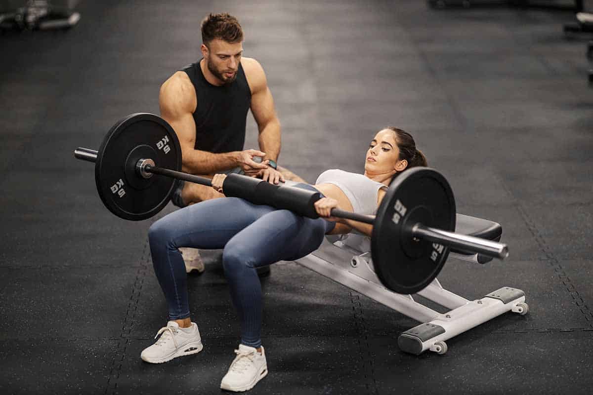 Strong female bodybuilder doing hip thrusts on bench at gym with her fitness instructor.