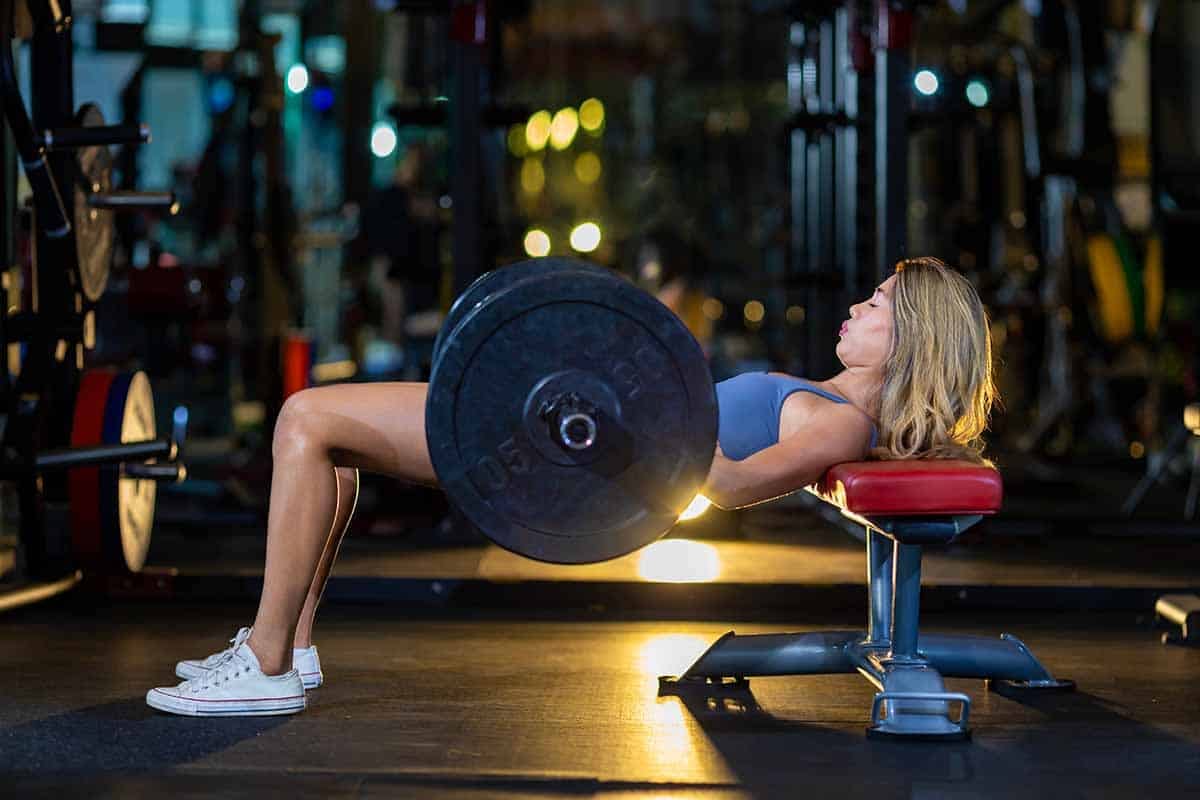 Asian woman is practice weight lifting hip thrusts position on barbell for core muscle inside gym with dark background for exercising and workout concept