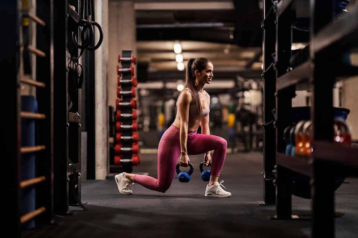 A fit female bodybuilder is doing lunges with kettlebells in a gym.