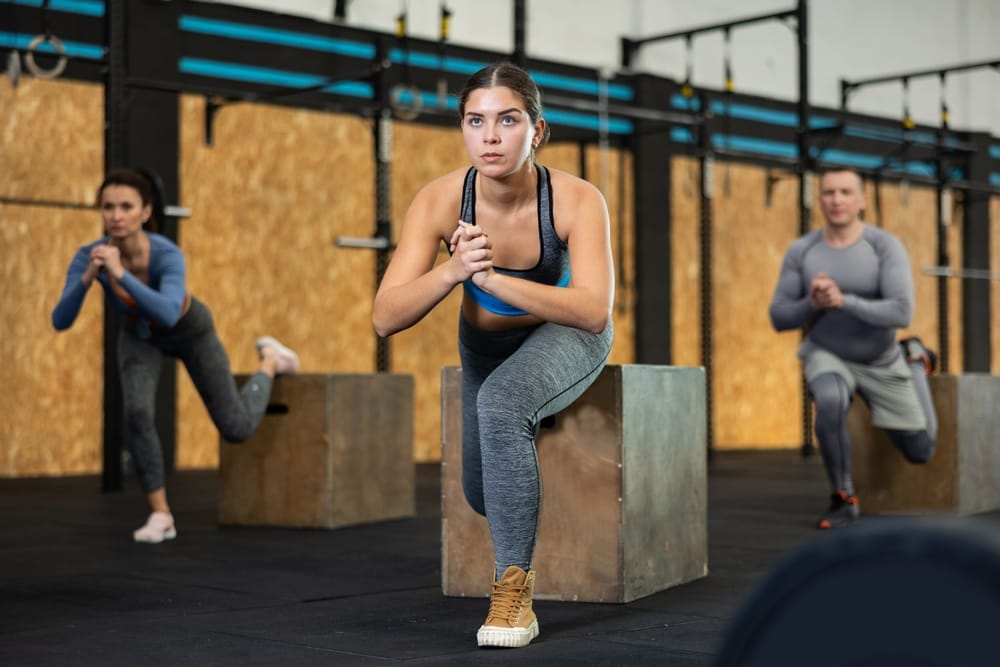 Concentrated young girl in fitness attire performing Bulgarian split squats using wooden plyometric box at gym.